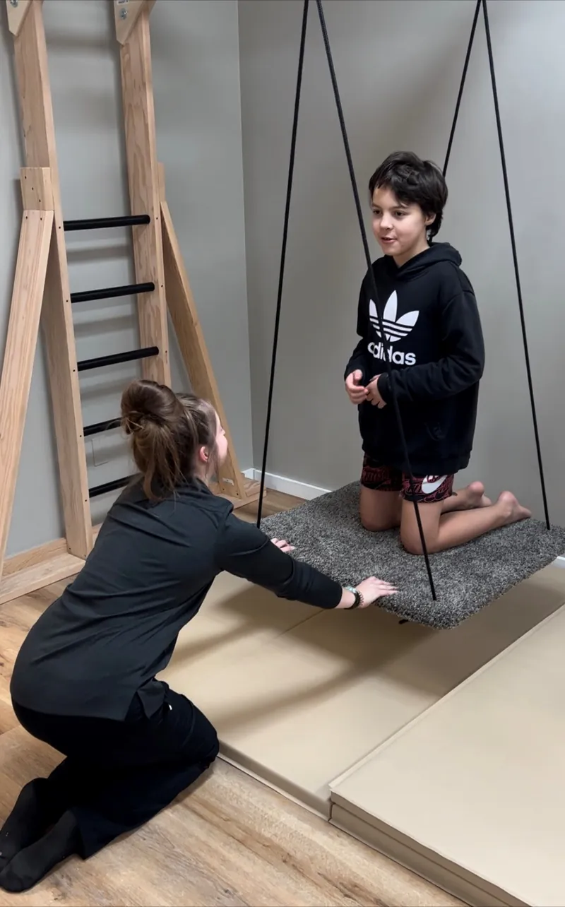 A BeKids therapist supporting a boy kneeling on a platform swing beside a climbing ladder in the therapy gym
